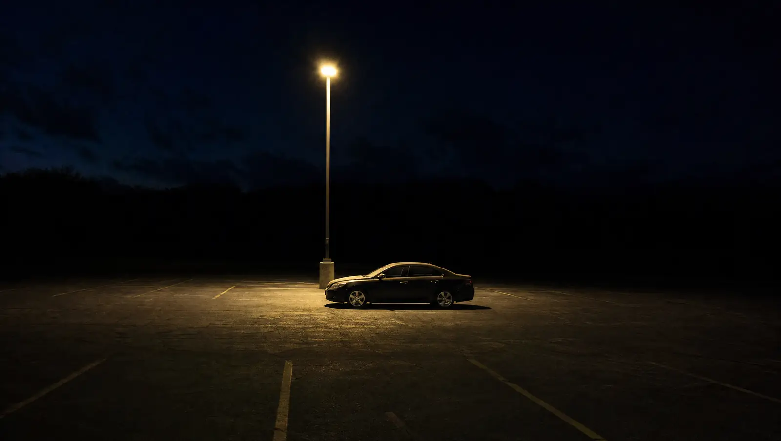 A single sedan sits alone in an empty parking lot at night under one street lamp.