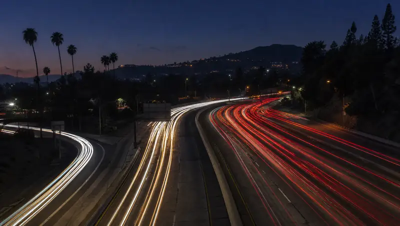 Headlight and tail-light trails converge where an on-ramp merges onto the 405 freeway at dusk.