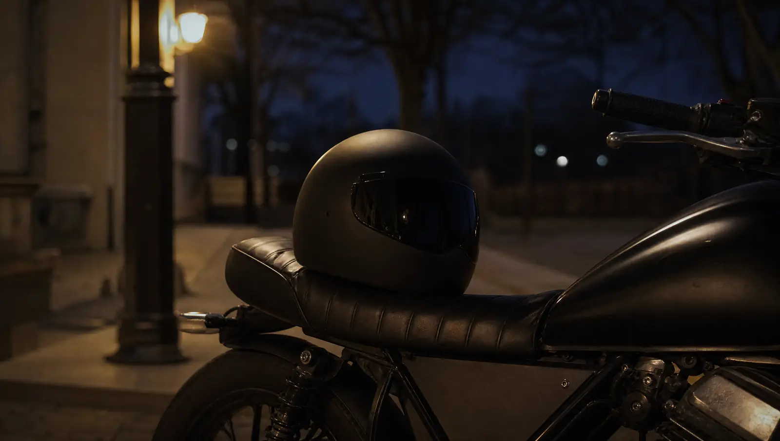 A black helmet rests on the seat of a parked motorcycle at dusk under a warm street lamp.