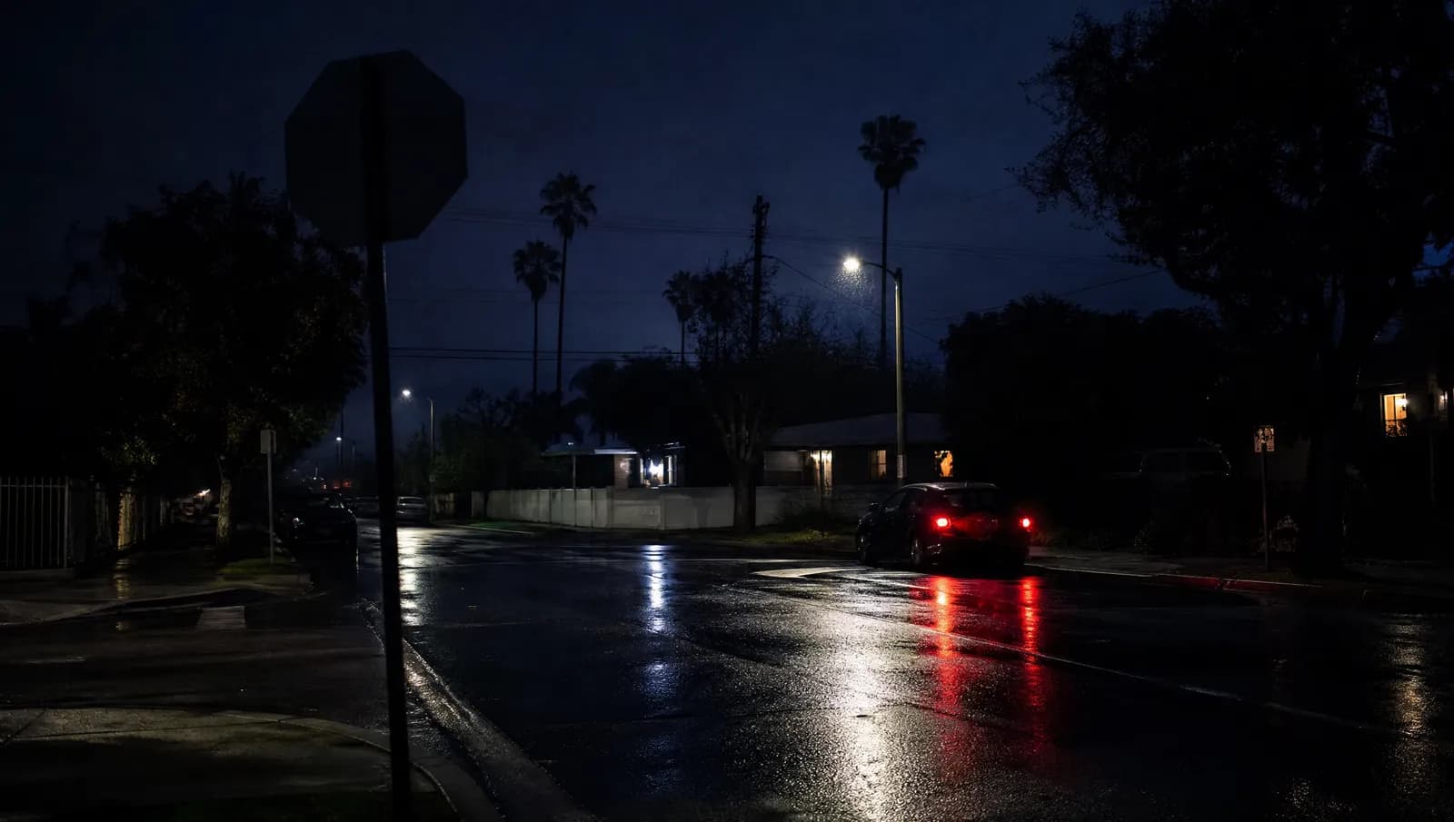 A rain-slicked Los Angeles residential intersection at night with a parked car's hazard lights blinking