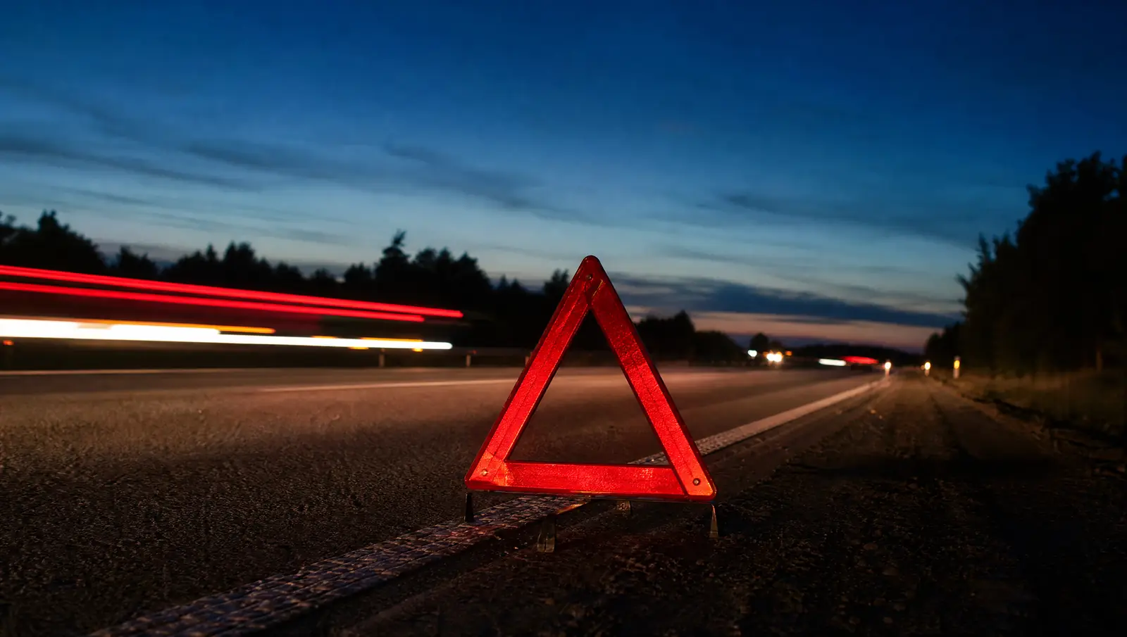 A red reflective hazard triangle stands on a freeway shoulder at dusk as headlights streak past.