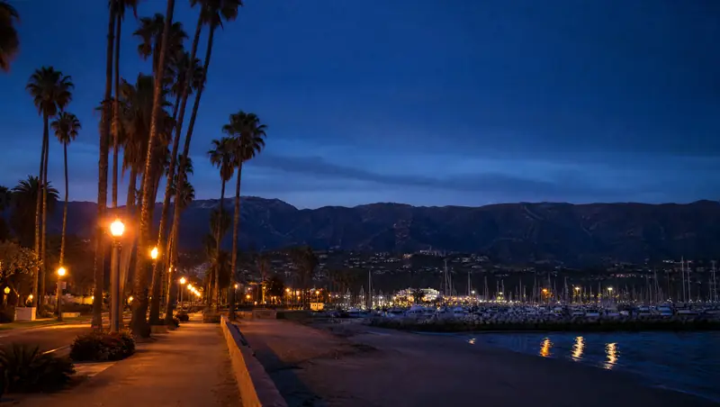 Palm trees and lamp posts line the Santa Barbara waterfront at dusk with the harbor and the Santa Ynez Mountains behind.