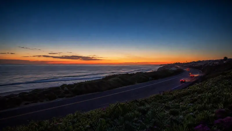 The coast highway curves along the Carlsbad bluffs at dusk with the Pacific Ocean glowing beyond.