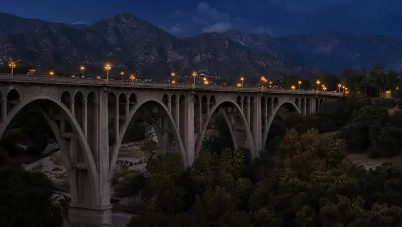 The arches of the Colorado Street Bridge in Pasadena glow with lamplight at dusk beneath the San Gabriel Mountains.