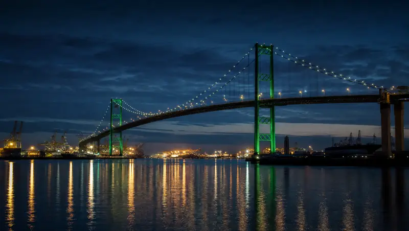 The Vincent Thomas Bridge is lit green over the Los Angeles harbor at blue hour with port cranes in the distance.